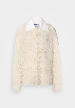 Krajková blůza s pevným bílým límcem, zdobená složitými květinovými vzory na krémovém materiálu, dlouhými rukávy a volným střihem.