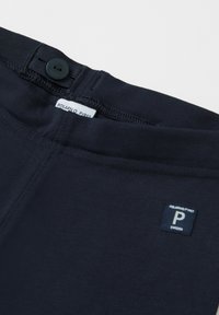 Tummansininen puuvillavyötärö, jossa on napitussuljenta ja ommellut yksityiskohdat, sisältäen lapun, jossa lukee "POLARN O. PYRET" sekä logotagin.