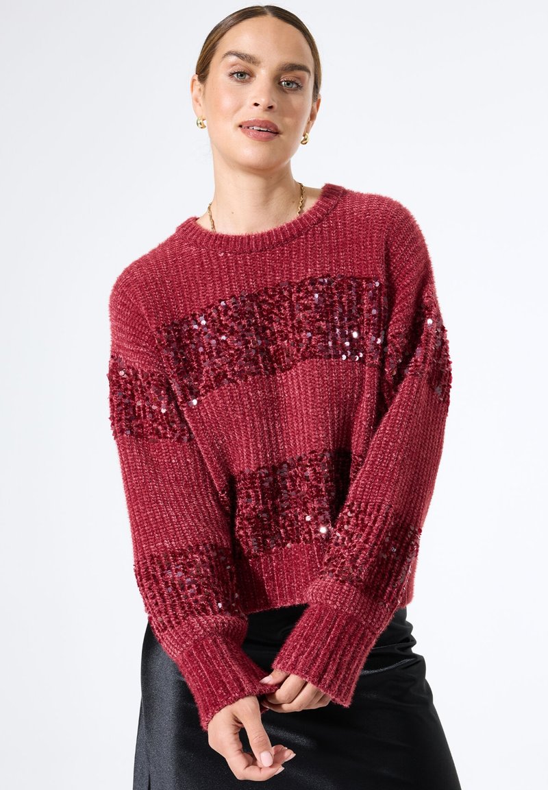 Vrouw in een rode grof gebreide trui met horizontale glanzende sequins en een zwarte satijnen rok, poserend tegen een witte achtergrond.