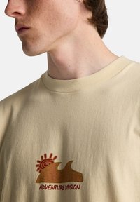 Μπεζ βαμβακερό μπλουζάκι με στρογγυλή λαιμόκοψη. Διαθέτει τυπωτό σχέδιο με ένα κύμα και ήλιο σε γήινους τόνους, με την επιγραφή "ADVENTURE DIVISION".