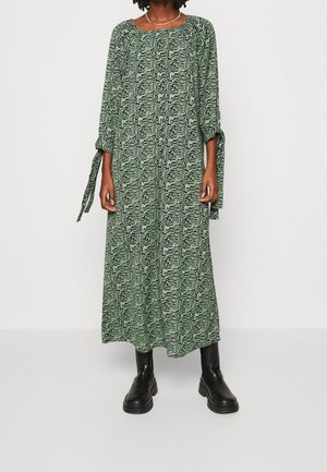 Robes longues et vertes avec un motif noir ondulé, un col rond et des manches longues avec des liens. Porté avec des bottines noires.
