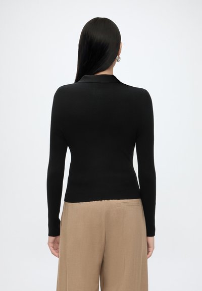 Femme aux longs cheveux noirs portant des boucles d’oreilles créoles argentées, un haut noir moulant à manches longues et un pantalon beige à jambes larges, vue de dos sur fond blanc.