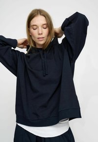Granatowy oversize'owy hoodie wykonany z lekkiego materiału, z wysokim kołnierzem, sznurkiem do regulacji i objętościowymi rękawami, nakładany na biały top.