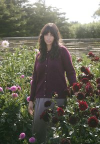 Mujer con cabello largo y oscuro que lleva un cárdigan color berenjena, de pie entre flores rosas y rojas oscuras en un jardín iluminado por el sol, con una cerca de madera detrás.