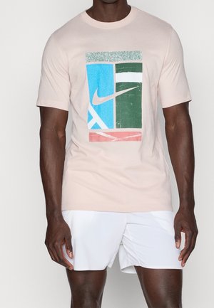 Mand iført en lysebeige T-shirt med et blåt, grønt og rødt grafikmotiv og hvide shorts, der står mod en ensfarvet baggrund.