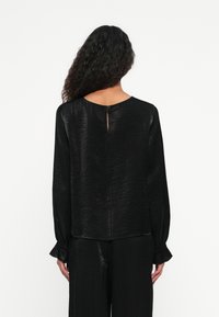 Schwarze Langarmbluse mit strukturiertem Finish, abgerundetem Saum und kleinem Schlüssellochöffnung im Rücken. Mit Rüschenmanschetten für zusätzliche Details.