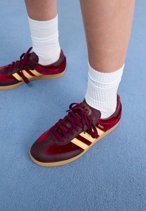 Person iført hvide crew-sokker og bordeaux Adidas Samba sneakers med gule striber på blå overflade.