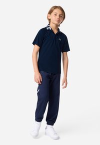 Ναυτικό πόλο μπλουζάκι με αντίθετο γιακά, συνδυασμένο με ναυτικά παντελόνια τζόγκινγκ και λευκά αθλητικά παπούτσια. Το σύνολο διαθέτει λογότυπο στη μπλούζα.