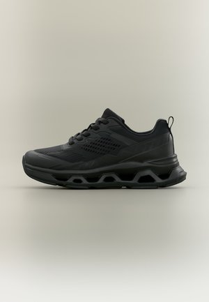 Zwarte atletische sneaker met mesh bovenwerk, dikke zool met geometrische uitsparingen, vetersluiting en hiel-lus op een neutrale achtergrond.