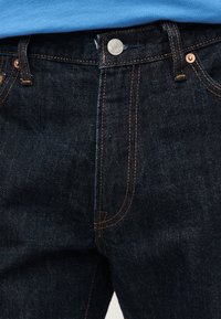 Σκούρο μπλε τζιν τζιν με χαλκομανίες, πορτοκαλί ραφές και ασημένιο κουμπί στη μέση κάτω από μπλε πουκάμισο.