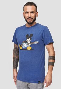 Blå bomulls t-shirt med en grafik av Mickey Mouse som spelar gitarr. Rund hals, kort ärm, med en etikett längst ner till vänster.