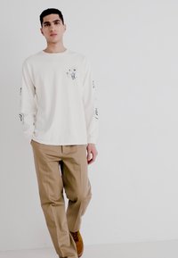 Camisa de manga larga color crema con gráfico frontal y diseños negros en las mangas, combinada con pantalones marrón claro y zapatos marrones.