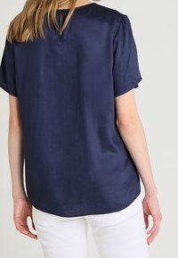 Kvinna med långt blont hår som bär en marint blå kortärmad blus och vita byxor, sedd bakifrån mot en enfärgad bakgrund.