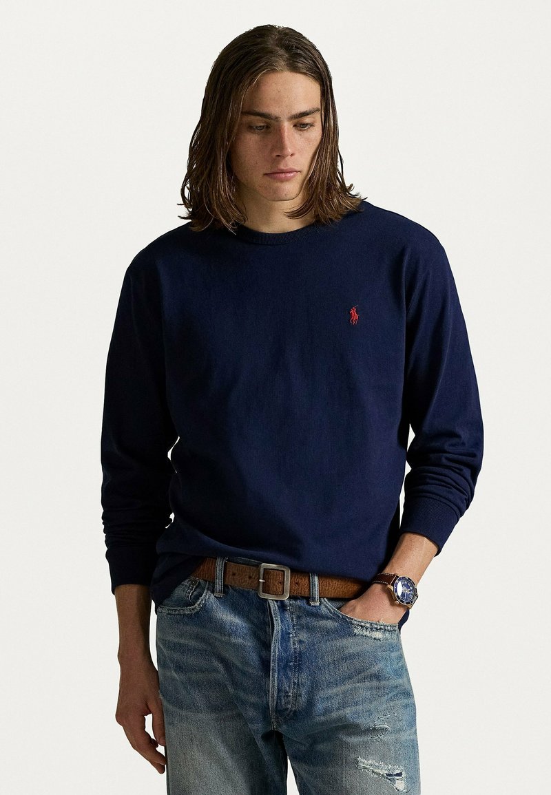 Camicia a maniche lunghe blu navy in tessuto morbido con un piccolo logo rosso sul petto, abbinata a jeans blu sbiaditi e una cintura marrone.