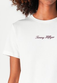 T-shirt en coton blanc avec des manches courtes et un col rond. Le nom de la marque "Tommy Hilfiger" est brodé en bordeaux sur le devant.
