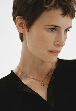 Mujer con pelo corto y castaño, que lleva una blusa negra y un delicado collar dorado con un colgante en forma de "G" en una cadena de cuentas.