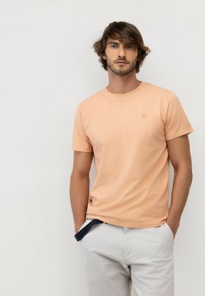 Hombre con cabello castaño medio que lleva una camiseta de manga corta color melocotón y pantalones gris claro, de pie con las manos en los bolsillos frente a un fondo liso.