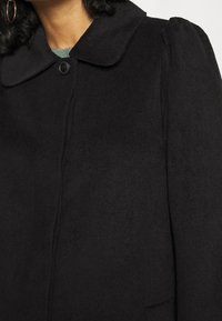 Femme portant un manteau en laine noir avec un col arrondi, des épaules bouffantes, un bouton visible, et un pull vert en dessous.