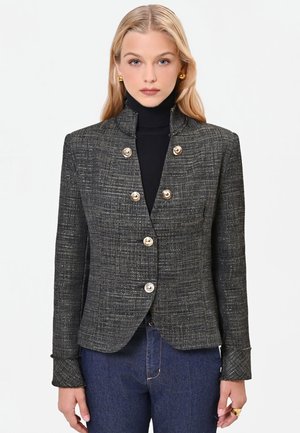 Donna bionda che indossa un blazer grigio scuro strutturato con bottoni dorati sopra un dolcevita nero e jeans blu, rivolta in avanti con espressione neutra.