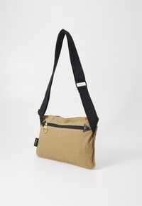 Torbka crossbody w kolorze brązowym z zamykaną na zamek kieszenią, czarnym paskiem i skórzanymi akcentami. Prostokątny kształt, gładka tekstura i minimalistyczny design.
