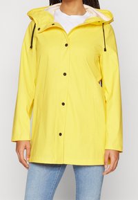 Veste de pluie imperméable jaune avec capuche, boutons-pression et deux poches latérales ; texture de tissu lisse ; accents de quincaillerie noirs contrastants.