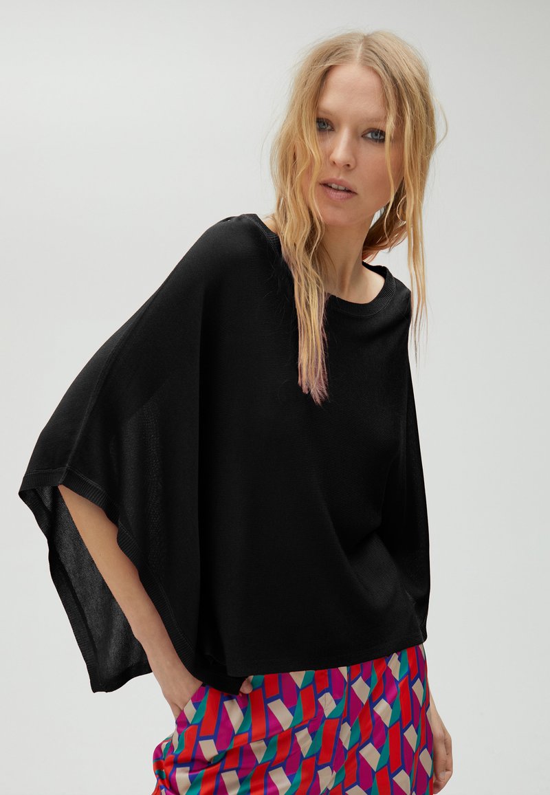 Černý volný top vyrobený z lehkého materiálu s širokými rukávy. Kombinován s barevnými kalhotami s geometrickými tvary.