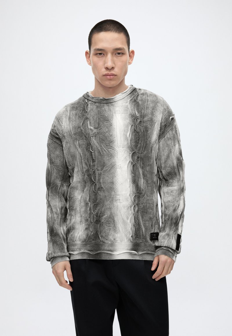 Jeune homme aux cheveux courts portant un pull en maille torsadée texturée gris et blanc et un pantalon noir, debout devant un fond uni.