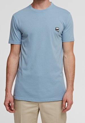 Hombre vistiendo una camiseta de manga corta azul claro con un pequeño logo en blanco y negro en el lado izquierdo del pecho, combinada con pantalones beige.