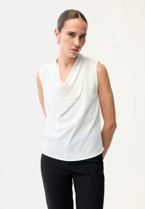 Kobieta z zaczesanymi do tyłu włosami, ubrana w białą, bezrękawnikową bluzkę z luźnym dekoltem oraz czarne spodnie, stoi na tle jednolitego białego tła.