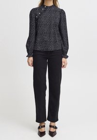 Blusa nera a pois con maniche lunghe, colletto alto e bottoni sul busto. Abbinata a pantaloni neri a gamba dritta e scarpe con punta.