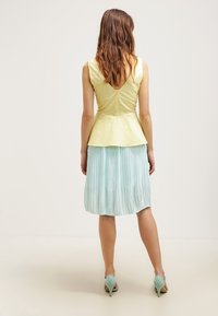 Top peplum in cotone giallo chiaro con chiusura a bottone sul retro, abbinato a una gonna plissettata verde menta. Le scarpe sono tacchi azzurri.