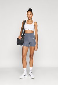 Pantaloni scurți atletici gri și un sutien sport alb cu logo-ul Adidas. Modelul poartă o geantă neagră de umăr și încălțăminte sport albă.