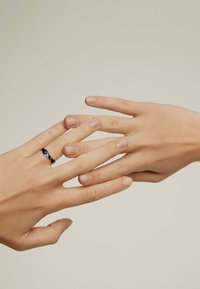 Anillo de banda de oro con acentos azules, llevado en una mano, mientras dos manos se tocan suavemente. Las uñas son naturales con un ligero esmalte. Fondo suave y neutro.