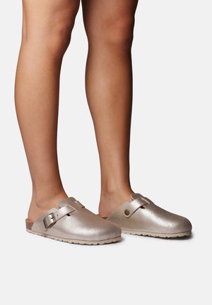 Metaliczne złote chodaki z teksturowaną powierzchnią, regulowaną klamrą i odkrytym tyłem, posiadające profilowaną wkładkę i antypoślizgową podeszwę.