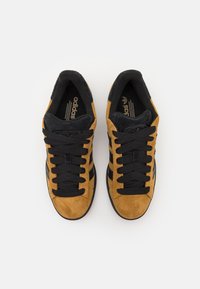 Ett par bruna mockaskor från Adidas med svarta ränder och svarta snören, sedd ovanifrån på en vit bakgrund.