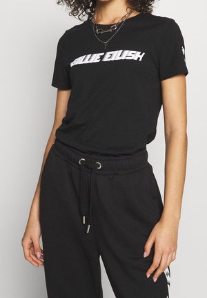 Kobieta w czarnym t-shircie Billie Eilish i czarnych spodniach z troczkami, ozdobiona warstwowymi naszyjnikami.