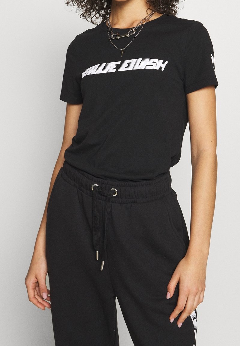 Femme portant un t-shirt noir Billie Eilish et un pantalon noir à cordon, accessoirisée avec des colliers superposés.