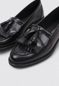 Mocasines de cuero negro con detalles de borlas, textura lisa, punta redonda, costuras en contraste y suela de goma de perfil bajo.