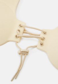 Bralette élastique beige avec une texture lisse, présentant un design à lacets à l'avant, des oeillets et un embout en plastique pour l'ajustement.