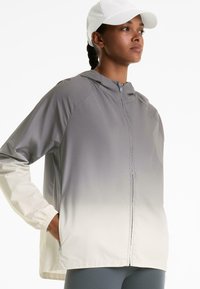Giacca leggera con cappuccio, sfumata dal grigio al crema. Presenta maniche a raglan e polsini elastici, realizzata in tessuto liscio e resistente all'acqua.