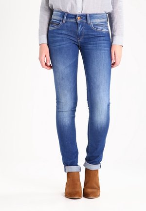 Frau trägt hochgekrempelte, schmale blaue Jeans, ein gestreiftes, in die Hose gestecktes Hemd und braune Wildleder-Stiefeletten, steht vor weißem Hintergrund.