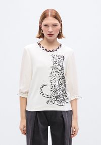 Λευκό τοπ με λεοπάρ σχέδιο, λαιμόκοψη στρογγυλή και λεπτομέρεια λεοπάρ. Διαφανή μανίκια με ελαστικά μανσέτα. Συνδυασμένο με σκουρόχρωμο παντελόνι με πιέτες.