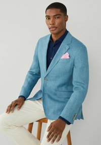 Blazer in lino azzurro chiaro con due tasche frontali, accompagnato da un fazzoletto rosa. Indossato sopra una camicia blu scuro e pantaloni beige.