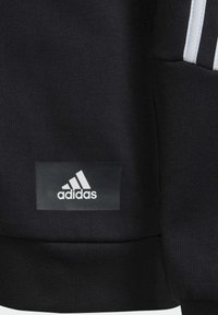 Sort sweatshirt lavet af struktureret stof, med hvide detaljer og et fremtrædende hvidt Adidas-logo på en rektangulær sort lap.