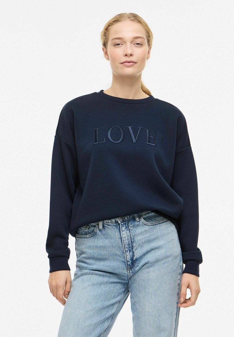 Sweat-shirt bleu marine avec "LOVE" en relief dans une teinte légèrement plus claire, featuring un col rond et des épaules tombantes, assorti à un jean bleu clair.