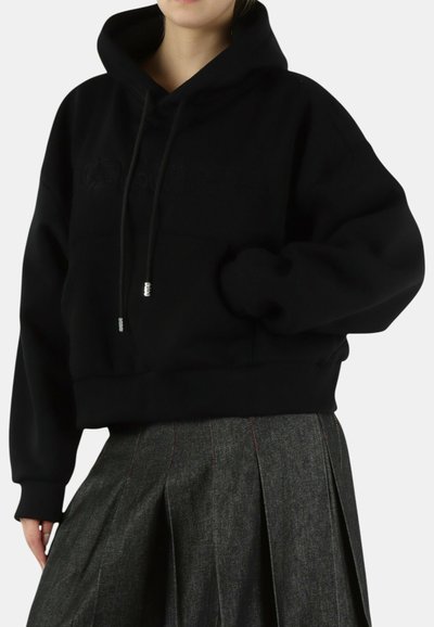Persoon draagt een zwart cropped hoodie met trekkoorden en een donker geplooide rok, hand in de zak van de hoodie gestoken.