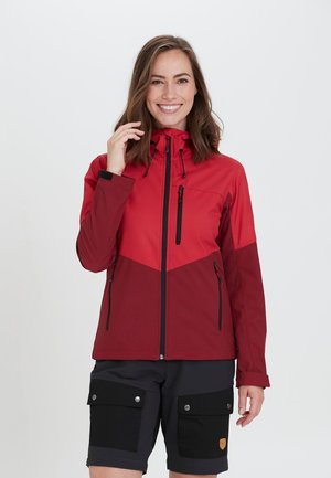 Red and burgundy jacket with a hood, featuring zippered pockets and a chevron design. Worn with black shorts and a smooth fabric texture.