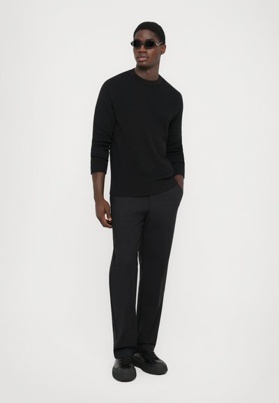 Zwarte geribbelde trui, zwarte op maat gemaakte broek en zwarte veterschoenen. Accessoires zijn smalle zwarte zonnebrillen. Soepele textuur, strak model.