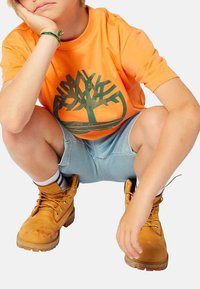 T-shirt arancione a maniche corte con logo di un albero verde, pantaloni corti in denim e stivali marroni con lacci, dalla finitura testurizzata e suole in gomma.