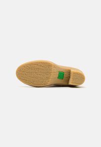 Suela de zapato de goma en beige, texturizada con surcos para un mejor agarre. Presenta un acento de logo verde cerca del talón, indicando la identidad de la marca.